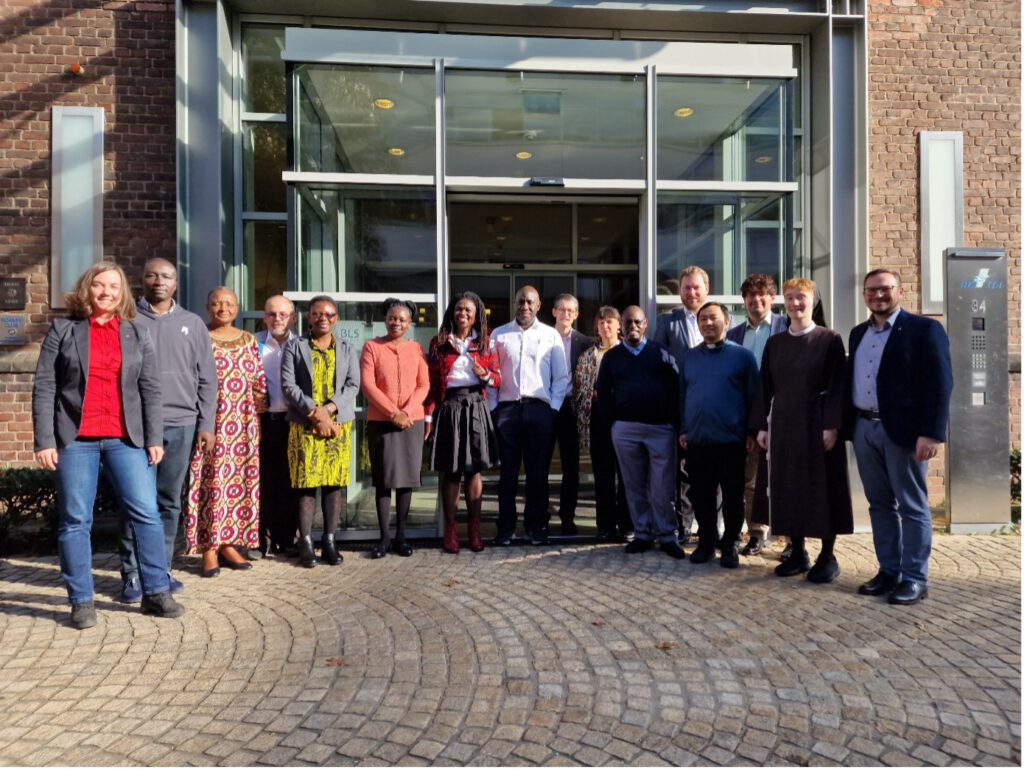 Gruppenfoto von etwa 18 Personen verschiedener Herkunft vor modernem Gebäude mit Glasfront und Backsteinwand.